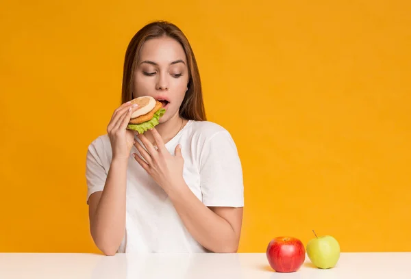 Menina com fome comer hambúrguer insalubre em vez de frutas de maçã — Fotografia de Stock