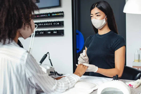 Salão de manicure e pedicure, conceito covid-19. Menina asiática em máscara protetora desinfecta unhas para o cliente — Fotografia de Stock