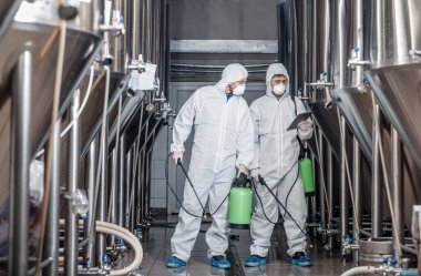 Guys in hazmat suits and masks disinfect brewery kettles at craft plant clipart