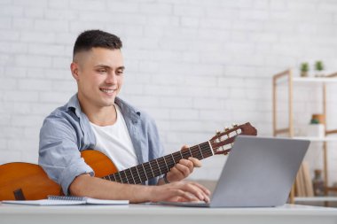 Covid-19 salgını sırasında online müzik dersi. Gitarlı gülümseyen genç adam dizüstüne bakıyor.