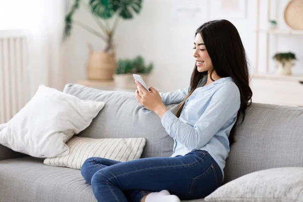 Descanso doméstico. Mujer asiática alegre que relaja en sofá con Smartphone, hojeando Internet — Foto de Stock