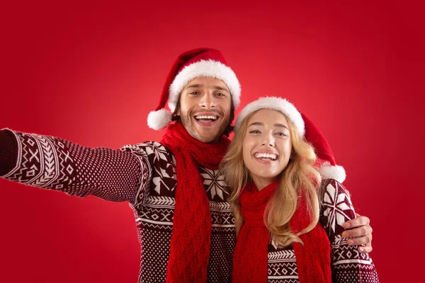Felice giovane ragazzo con la sua ragazza in abiti di Natale prendendo selfie su sfondo rosso — Foto Stock