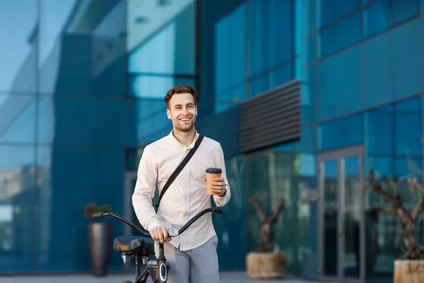 Szczęśliwy facet z kawą na wynos i rowerem na tle biurowca idzie do pracy — Zdjęcie stockowe