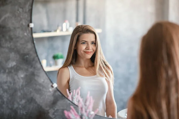 Приваблива молода леді з сяючою шкірою і шовковистим довгим волоссям дивиться на її відображення в дзеркалі у ванній — стокове фото