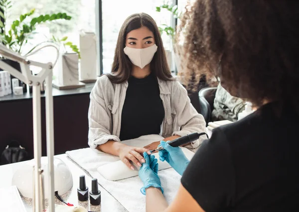 Work of manicure and pedicure salon. Asian girl looks at master in protective mask during electrical nail procedure