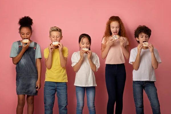 Adorables enfants multiethniques mangeant de délicieux beignets sucrés sur fond rose — Photo