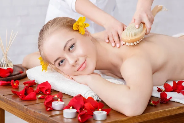 Masaje con cepillo. Sonriente chica recibe masaje en mesa de masaje con pétalos de rosa —  Fotos de Stock