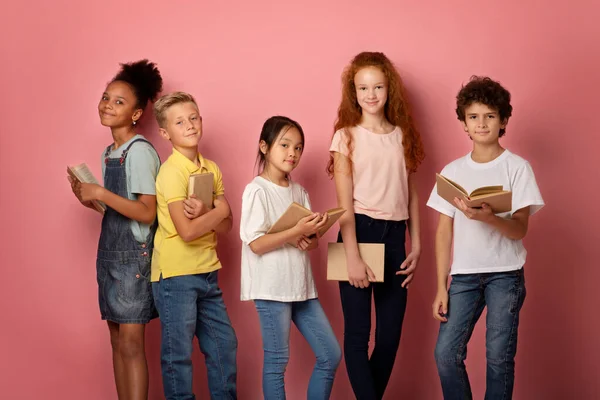 Escolares multinacionais com livros didáticos olhando para a câmera sobre fundo rosa, retrato — Fotografia de Stock