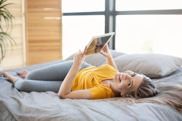 Hermosa joven viendo webinar o película en la computadora tableta en su cama en casa — Foto de Stock