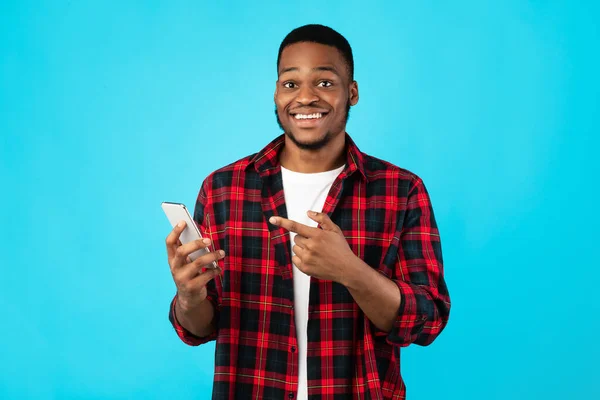Afrykański facet wskazując palec na smartfona stojącego nad niebieskim tle — Zdjęcie stockowe