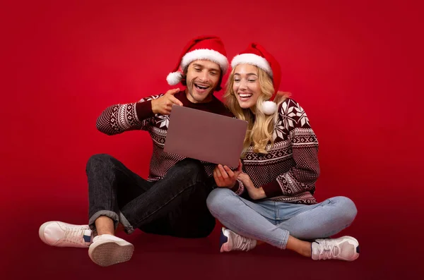 Kırmızı arkaplandaki dizüstü bilgisayarı gösteren Noel elbiseli genç bir çiftin tam boy portresi. — Stok fotoğraf