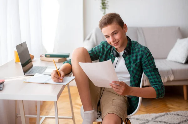 Fröhlicher Teenager macht seine Hausaufgaben, sitzt am Schreibtisch, hält Papiere in der Hand — Stockfoto