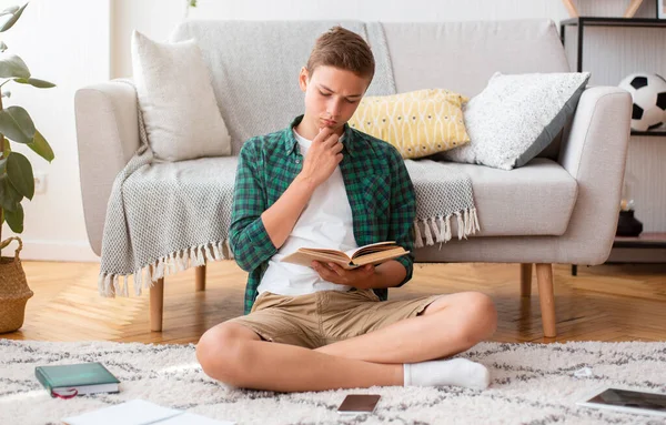 Konzentrierter Teenie-Junge sitzt auf dem Fußboden im Zimmer und liest Buch — Stockfoto