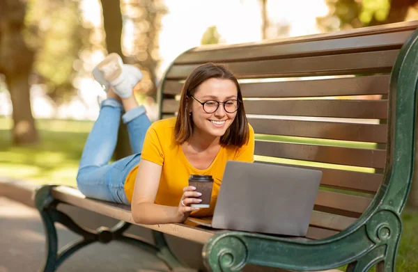 Estudiante chica usando ordenador portátil acostado en el banco en el parque — Foto de Stock