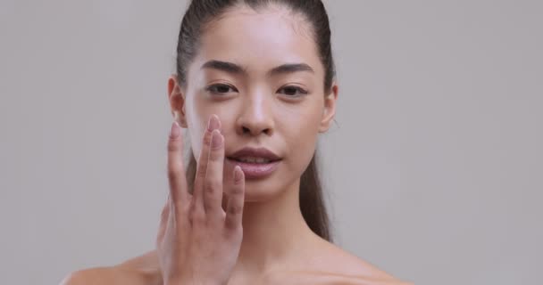 Ritratto di giovane donna asiatica che applica crema spalmare sulla guancia e sorridente alla macchina fotografica — Video Stock