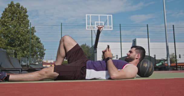 Баскетболист лежит на полу и просматривает на смартфоне, на открытом фоне корта — стоковое видео