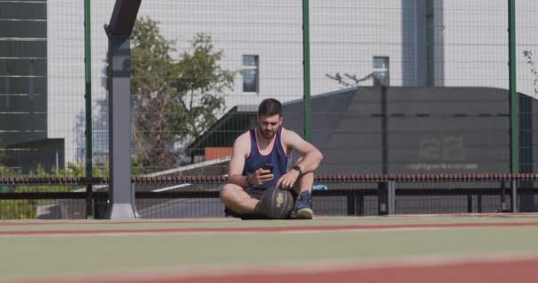 Молодой баскетболист ждет команду, болтает по мобильному телефону — стоковое видео