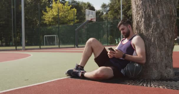 Utomhus porträtt av ung skäggig idrottsman med mobiltelefon på utomhus basketplan — Stockvideo