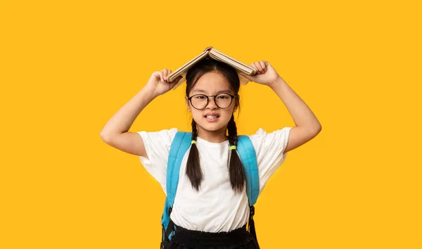 Elégedetlen ázsiai iskoláslány pózol könyv a fej a stúdióban — Stock Fotó