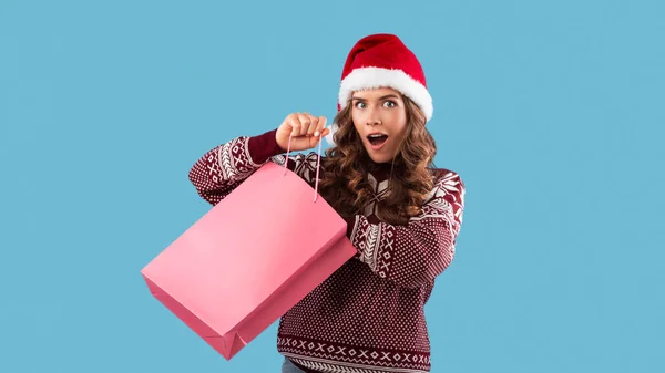 Señora sorprendida en sombrero de Santa y suéter de Navidad mirando en el bolso de compras sobre fondo azul, espacio para el diseño — Foto de Stock