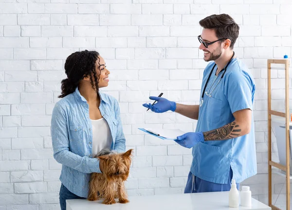 Молодой ветеринар-мужчина разговаривает с клиентом с маленькой собачкой в медицинском кабинете — стоковое фото