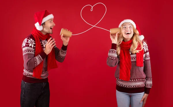 Christmas love message. Cute couple in Santa hats and sweaters talking on tin phone on red background