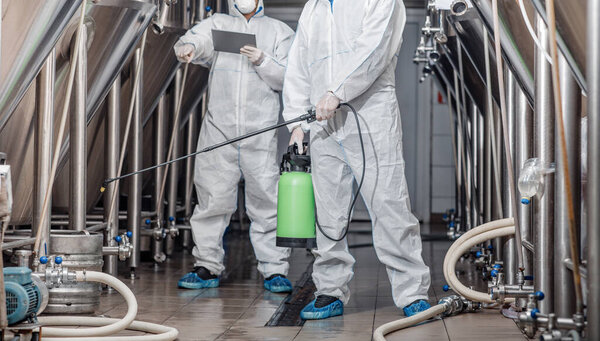 Workers in protective suits with spray bag and tablet in factory
