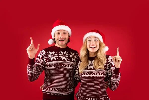 Alegre pareja milenaria en suéteres de lana y sombreros de Santa Claus apuntando hacia arriba sobre fondo rojo — Foto de Stock