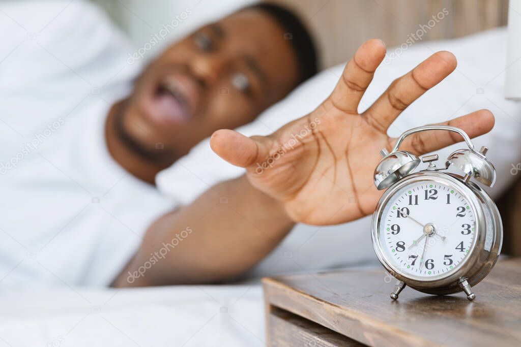 Stressed African Man Turning Off Alarm-Clock Lying In Bed Indoors