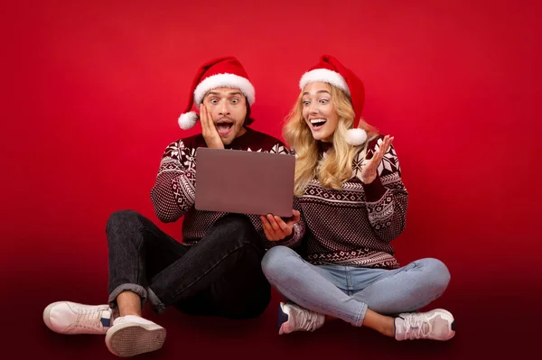 Young couple excited over Christmas discounts sitting with laptop on red background — Stock Photo, Image