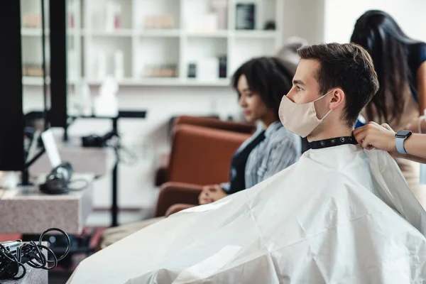 Hombre joven con máscara protectora sentado en la silla del cliente en la peluquería, esperando un corte de pelo —  Fotos de Stock