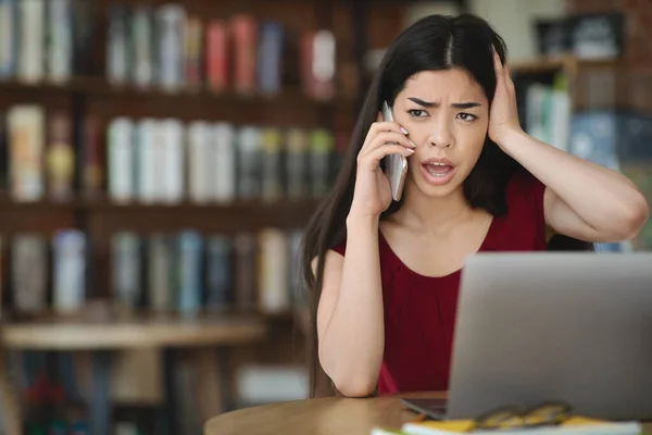 Frustrated asian woman having computer problems, talking on cellphone with customer support