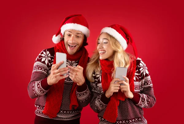 Christmas sale. Excited young couple in Santa hats shopping online on smartphones over red background