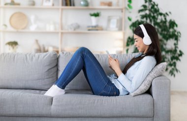 Akıllı telefonuyla koltuğa uzanmış, kulaklıkla müzik dinleyen Asyalı bir kız.