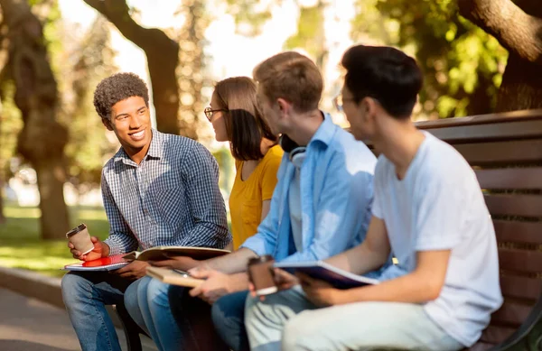 Cuatro estudiantes de secundaria discutiendo clases sentados en el banco en el parque — Foto de Stock