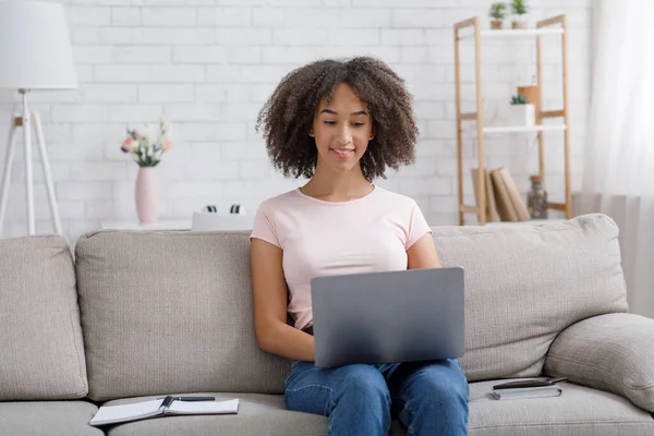 Vänlig afrikansk amerikansk flicka arbetar i laptop, sitter på soffa med anteckningsblock i vardagsrummet interiör — Stockfoto