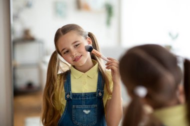 Makyaj yapan, elinde fırçayla aynaya bakan genç bir kız.