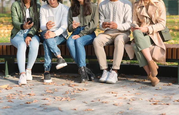 Przycięte z młodych przyjaciół za pomocą telefonów komórkowych, siedzi na ławce — Zdjęcie stockowe