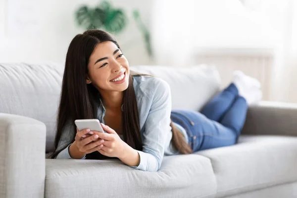 Unbekümmerte Asiatin genießt Ruhe auf bequemem Sofa mit Smartphone, mit freundlichem Lächeln — Stockfoto