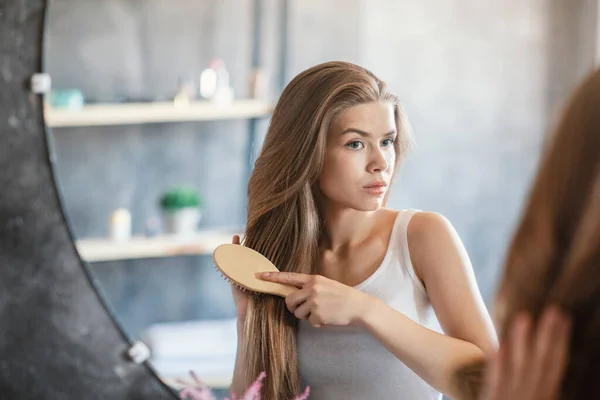 Menina encantadora escovando seu longo cabelo bonito na frente do espelho em casa — Fotografia de Stock