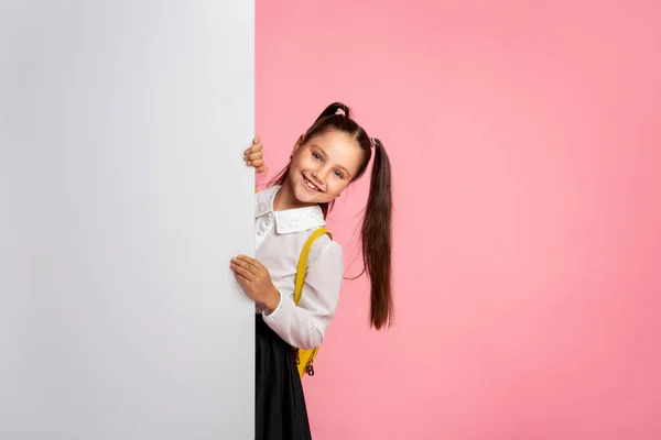 Készen állok a leckére. Vidám, egyenruhás iskoláslány néz ki fehér hirdetőtáblával. — Stock Fotó