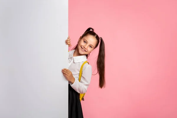 Listo para la lección. alegre colegiala en uniforme peeps fuera de con blanco cartel — Foto de Stock