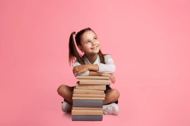 Neşeli liseli kız hayal kuruyor, kitap yığınında oturuyor ve boş bir yere bakıyor.
