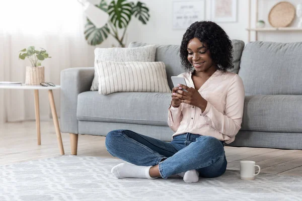 Mulher Africana Navegando nas mídias sociais no smartphone e bebendo café em casa — Fotografia de Stock