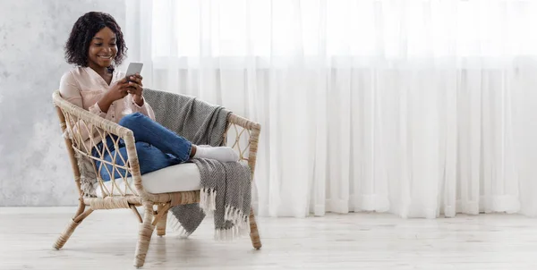 Χαλαρωμένο μαύρο κορίτσι κάθεται σε ψάθινη καρέκλα και τα μηνύματα στο Smartphone, Πανόραμα — Φωτογραφία Αρχείου