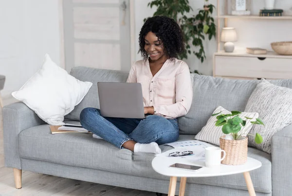 Op afstand werken. Vreugdevolle zwarte vrouw werken op laptop op bank thuis — Stockfoto