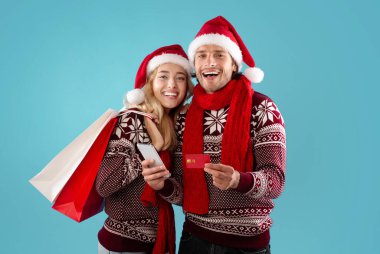 Romantik genç çift Noel hediyesi alışverişi yapıyor. İnternet üzerinden kredi kartı ve mavi arka planda akıllı telefon kullanıyorlar.