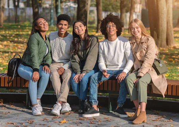 Grupo de amigos positivos sentados en el banco y sonriendo —  Fotos de Stock