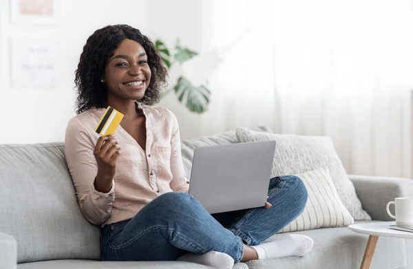 Compras en línea. Chica africana alegre posando en casa con el ordenador portátil y la tarjeta —  Fotos de Stock