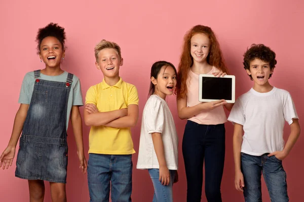 Szczęśliwy zróżnicowany uczniów gospodarstwa tablet z pustym ekranem na różowym tle, makieta do projektowania — Zdjęcie stockowe